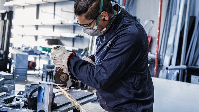 Seseorang yang mengenakan peralatan keselamatan menggiling logam, sehingga menimbulkan percikan api di bengkel.