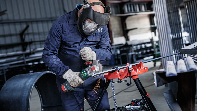 Seseorang yang mengenakan peralatan keselamatan menggunakan penggiling sudut untuk memotong logam, menghasilkan percikan api.