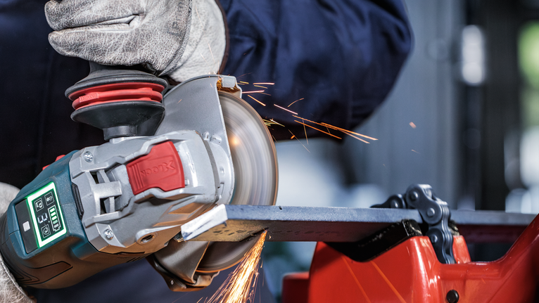 Seseorang yang mengenakan peralatan keselamatan menggiling logam dengan penggiling sudut, sehingga menimbulkan percikan api.