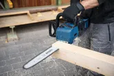 Seseorang yang mengenakan peralatan keselamatan memotong balok kayu dengan gergaji mesin tanpa kabel.