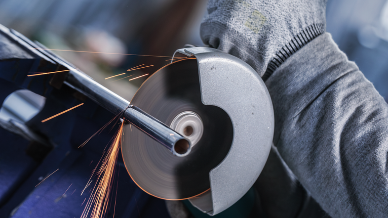Orang yang mengenakan peralatan keselamatan sedang memotong pipa logam dengan alat penggiling.