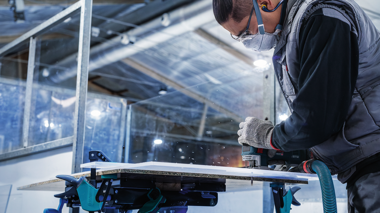 Seseorang yang mengenakan perlengkapan keselamatan menggunakan alat listrik untuk memotong panel kayu.