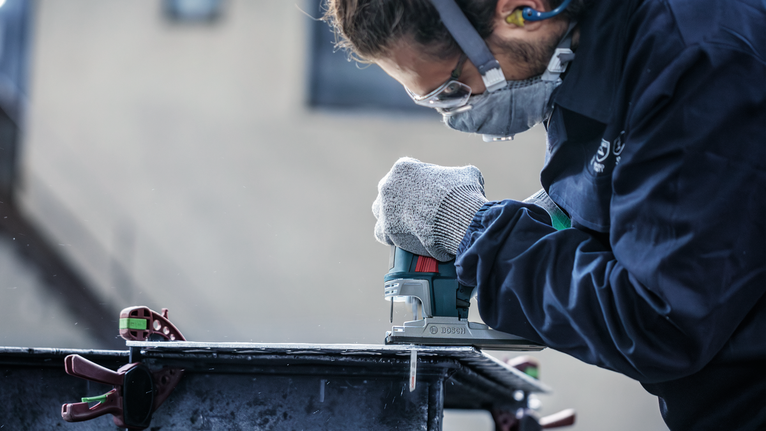 Orang yang mengenakan perlengkapan keselamatan menggunakan gergaji ukir untuk memotong lembaran logam.
