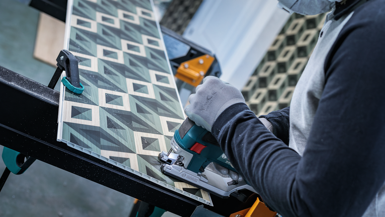 Orang yang mengenakan peralatan keselamatan memotong papan berpola dengan gergaji genggam.