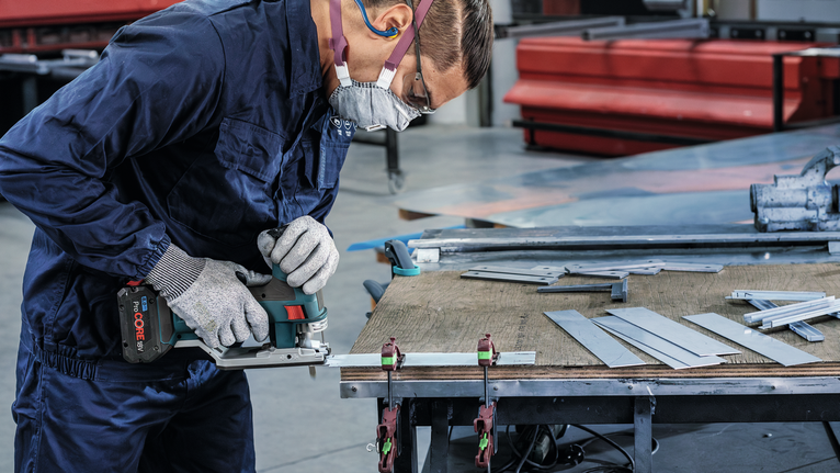 Seseorang yang mengenakan peralatan keselamatan memotong lembaran logam dengan gergaji listrik di meja kerja.