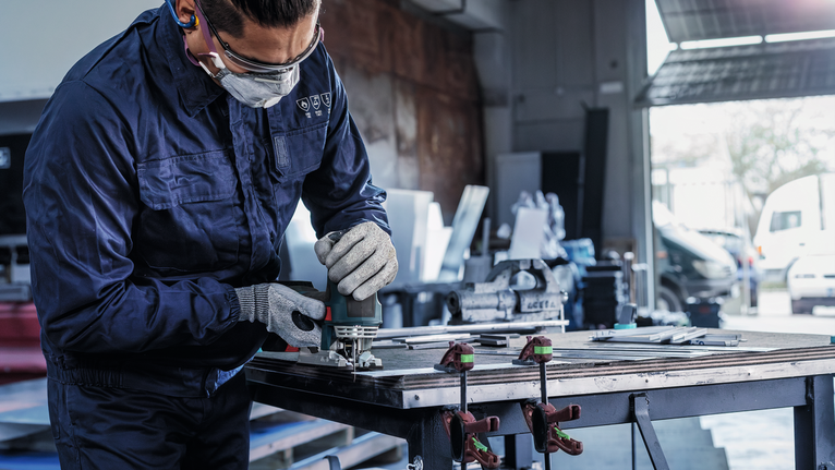 Orang yang mengenakan peralatan keselamatan menggunakan alat listrik untuk mengampelas logam di meja kerja.