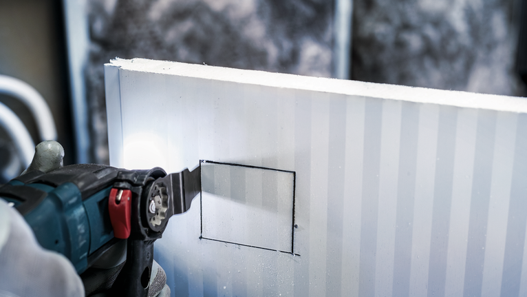 Seseorang yang mengenakan peralatan keselamatan sedang memotong lubang persegi di dinding kering dengan perkakas listrik.