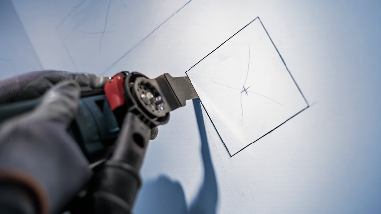 Orang yang mengenakan perlengkapan keselamatan menggunakan perkakas listrik untuk memotong persegi pada dinding kering.