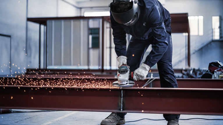 Seseorang yang mengenakan peralatan keselamatan menggiling balok logam, sehingga menghasilkan percikan api.