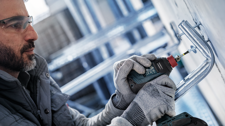 Seseorang yang mengenakan perlengkapan keselamatan menggunakan bor listrik untuk mengencangkan braket logam ke dinding.