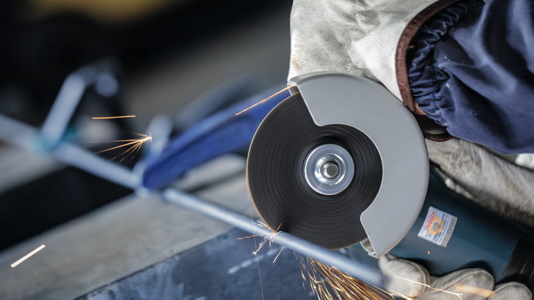 Orang yang mengenakan peralatan keselamatan menggiling batang logam dengan alat pemotong.