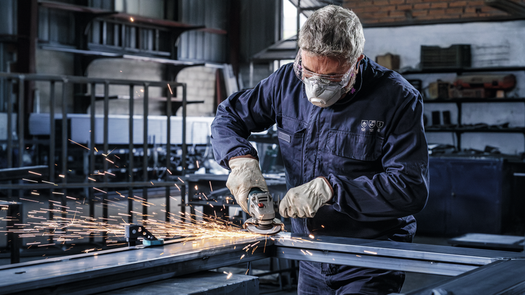 Seorang pekerja yang mengenakan peralatan keselamatan menggiling logam, menciptakan percikan api di sebuah pabrik.