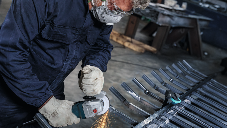 Orang yang mengenakan peralatan keselamatan menggiling batang logam dengan perkakas listrik.