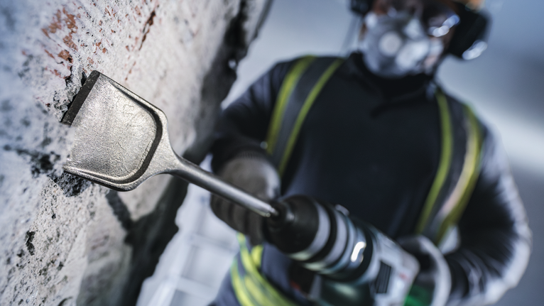 Orang yang mengenakan peralatan keselamatan menggunakan palu putar untuk memahat dinding beton.