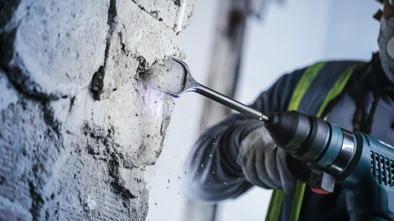 Seseorang yang mengenakan perlengkapan keselamatan menggunakan perkakas listrik untuk menghancurkan beton.
