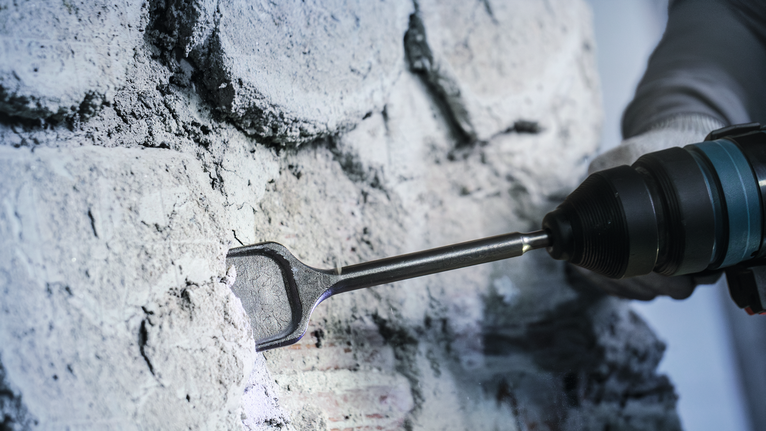 Orang yang mengenakan peralatan keselamatan sedang memahat dinding beton dengan alat listrik.