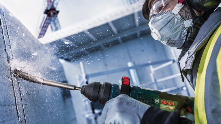 Chisel PRO SDS plus Hollow Gouging, 22 x 250 mm Seseorang yang mengenakan peralatan keselamatan mengebor beton, sehingga menimbulkan puing-puing yang beterbangan.