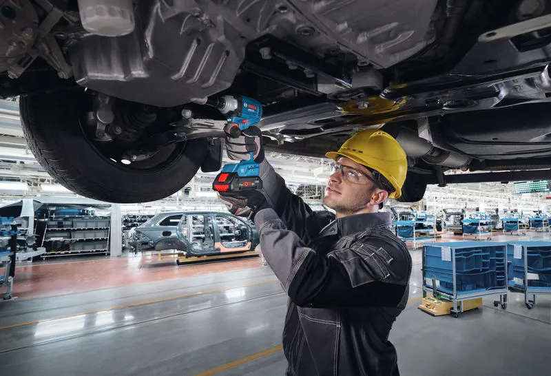 Seseorang yang mengenakan perlengkapan keselamatan menggunakan kunci pas listrik untuk mengencangkan baut di bawah rangka kendaraan.