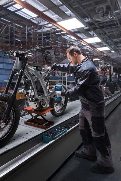 Seseorang yang mengenakan perlengkapan keselamatan menggunakan driver benturan untuk merakit rangka sepeda motor.