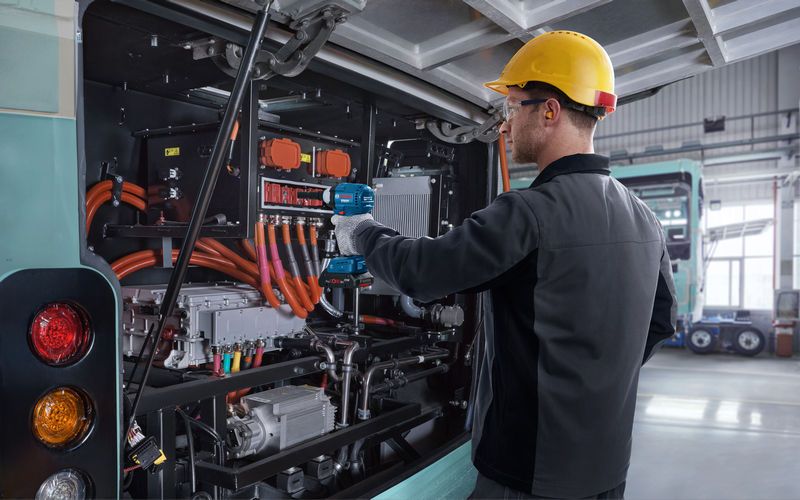 Seseorang yang mengenakan peralatan keselamatan menggunakan kunci benturan pada mesin bus listrik.