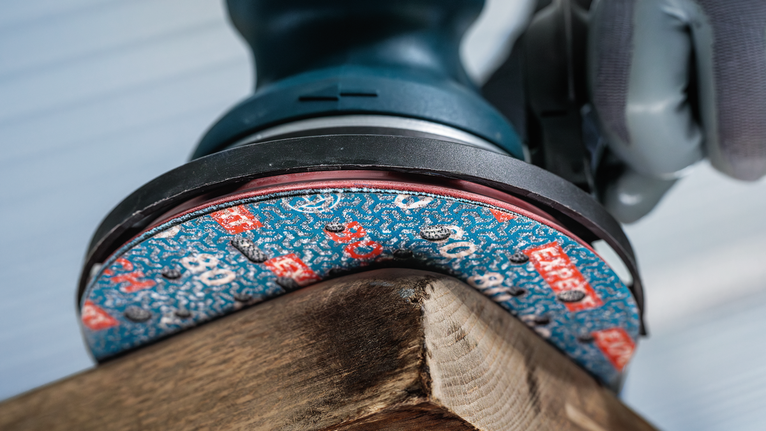 Orang yang mengenakan peralatan keselamatan sedang mengampelas sudut papan kayu.