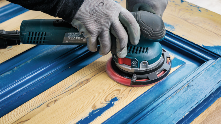 Seseorang yang mengenakan perlengkapan keselamatan mengampelas cat biru dari pintu kayu.