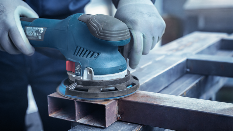 Seseorang yang mengenakan perlengkapan keselamatan mengampelas permukaan logam dengan mesin pengamplas.