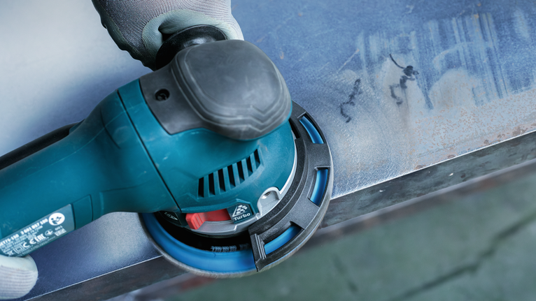 Orang yang mengenakan peralatan keselamatan sedang mengampelas permukaan logam dengan perkakas listrik.