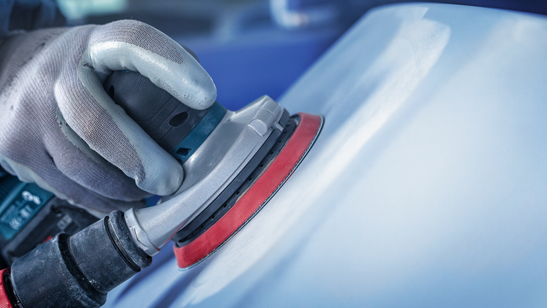 Orang yang mengenakan peralatan keselamatan sedang mengampelas permukaan mobil dengan mesin pengamplas.