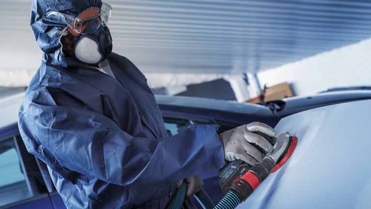 Seseorang yang mengenakan perlengkapan keselamatan mengampelas permukaan mobil dengan mesin pengamplas.
