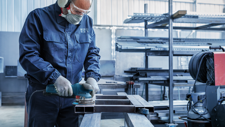 Seseorang yang mengenakan peralatan keselamatan sedang menggiling balok logam di bengkel industri.