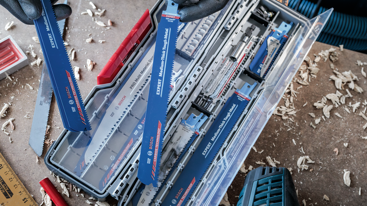 Pekerja bersarung tangan sedang menata mata gergaji biru dalam kotak plastik, dikelilingi serutan kayu.