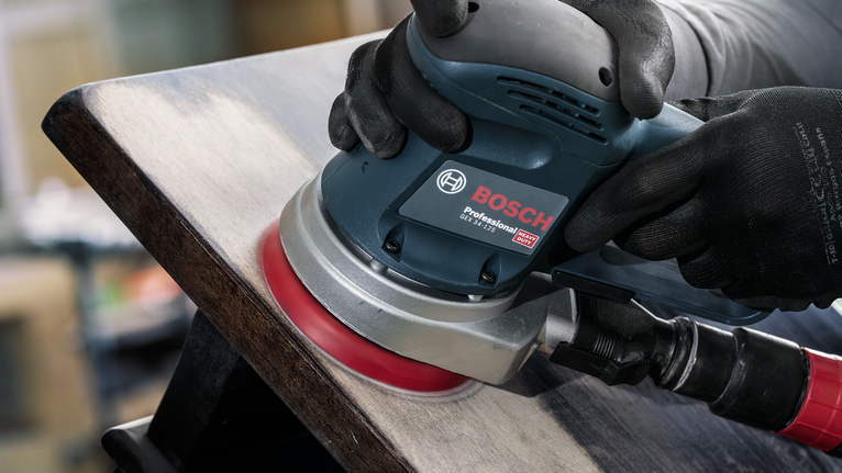 Orang yang mengenakan perlengkapan keselamatan mengampelas permukaan kayu dengan mesin pengamplas.