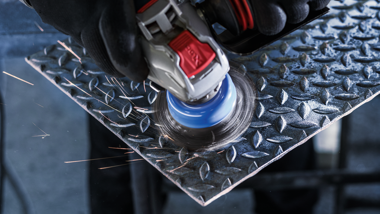 Sikat Mangkuk PRO Stainless Steel heavy X-Lock, 75 x 0,5 mm Orang yang mengenakan peralatan keselamatan menggiling lembaran logam saat percikan api beterbangan.
