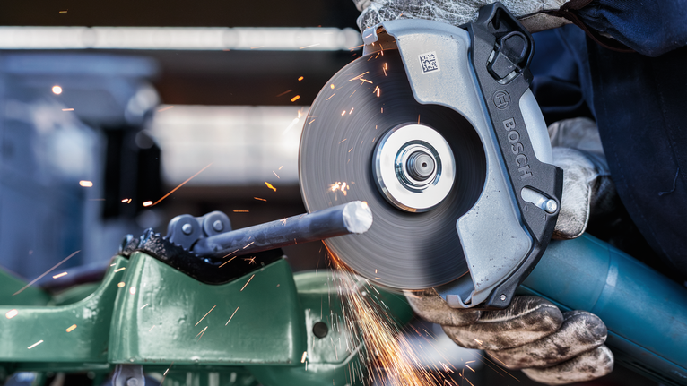 Seseorang yang mengenakan peralatan keselamatan memotong batang logam dengan penggiling sudut yang berputar.