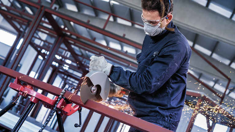 Seseorang yang mengenakan peralatan keselamatan menggunakan pemotong listrik untuk memotong pipa logam, yang menghasilkan percikan api.