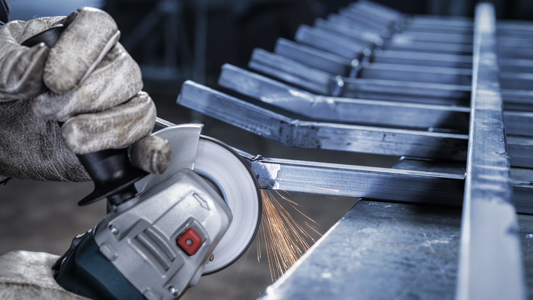 Orang yang mengenakan peralatan keselamatan menggiling batang logam dengan penggiling sudut, sehingga menimbulkan percikan api.