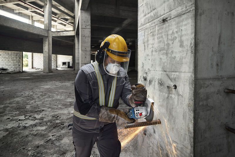Seseorang yang mengenakan peralatan keselamatan sedang mengasah tulangan beton pada dinding beton dengan penggiling sudut tanpa kabel.