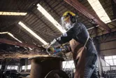 Seseorang yang mengenakan peralatan keselamatan menggunakan penggiling sudut tanpa kabel untuk memotong logam, yang menimbulkan percikan api.