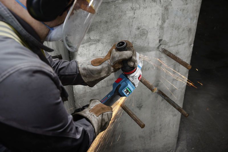 Seseorang yang mengenakan perlengkapan keselamatan menggiling tulangan beton yang terbuka pada dinding beton.