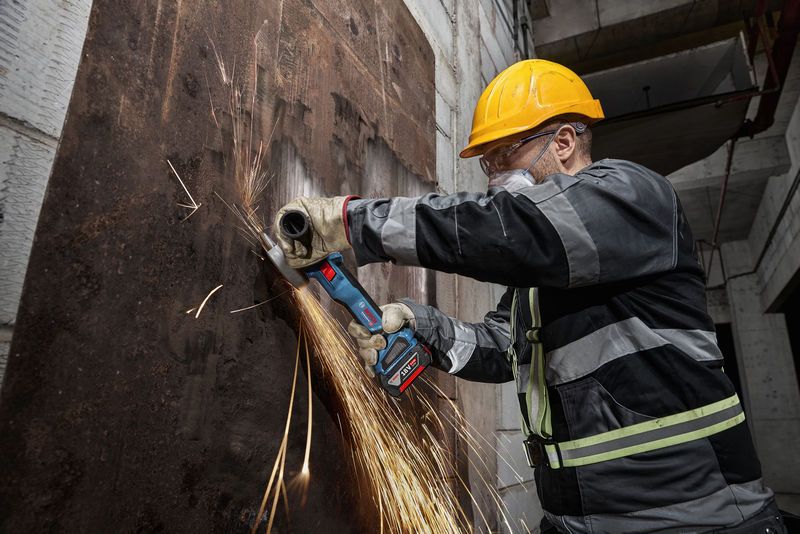 Seseorang yang mengenakan peralatan keselamatan menggiling lembaran logam berkarat dengan penggiling sudut tanpa kabel.