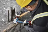 Seseorang yang mengenakan peralatan keselamatan menggiling tulangan beton pada beton dengan penggiling sudut tanpa kabel.