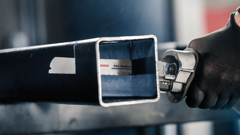 Orang yang mengenakan peralatan keselamatan memotong tabung logam persegi dengan gergaji bolak-balik.