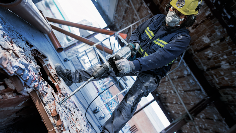 Seseorang yang mengenakan perlengkapan keselamatan menggunakan palu putar untuk memecahkan puing beton.