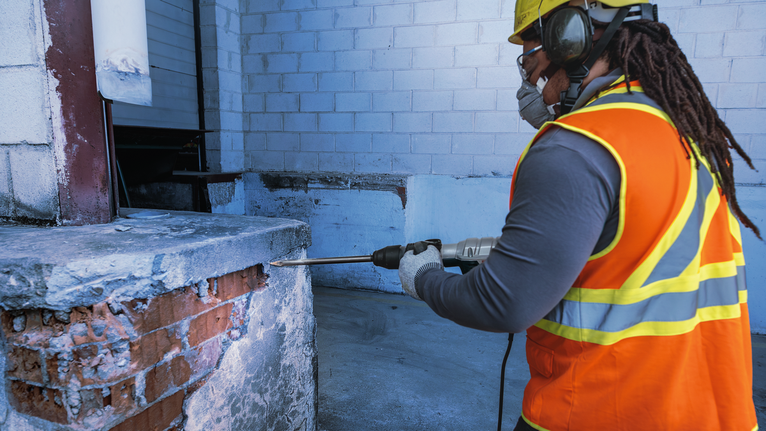 Seseorang yang mengenakan peralatan keselamatan mengebor dinding beton di lokasi konstruksi.
