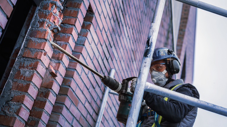 Seseorang yang mengenakan peralatan keselamatan mengebor dinding bata di perancah.