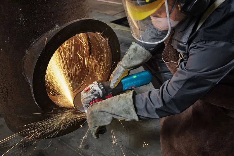 Seseorang yang mengenakan peralatan keselamatan menggiling pipa logam besar, sehingga menghasilkan percikan terang.
