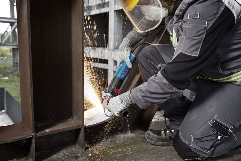 Seseorang yang mengenakan perlengkapan keselamatan sedang mengasah balok logam dengan penggiling sudut.