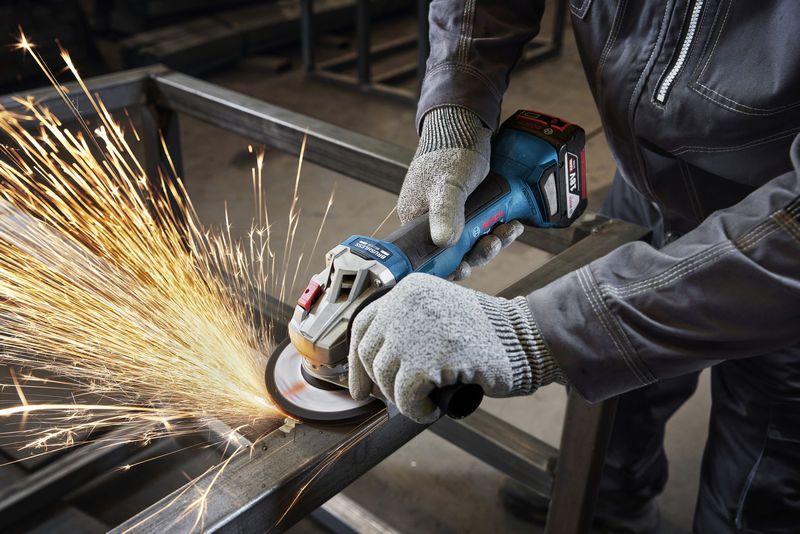 Seseorang yang mengenakan perlengkapan keselamatan sedang mengasah balok logam dengan penggiling sudut.