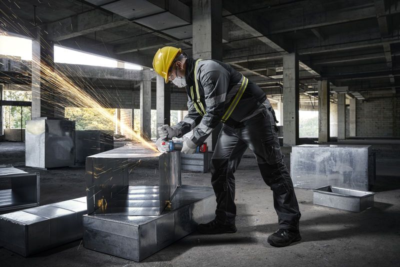 Seseorang yang mengenakan peralatan keselamatan menggunakan penggiling sudut pada saluran logam di lokasi konstruksi.